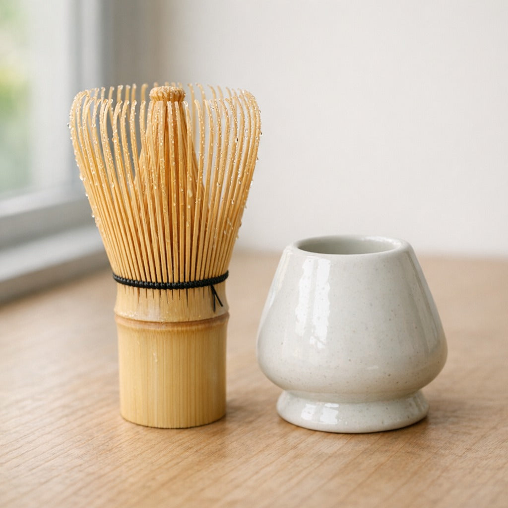 A freshly rinsed bamboo whisk with water droplets standing beside a white ceramic whisk holder on a light wood surface