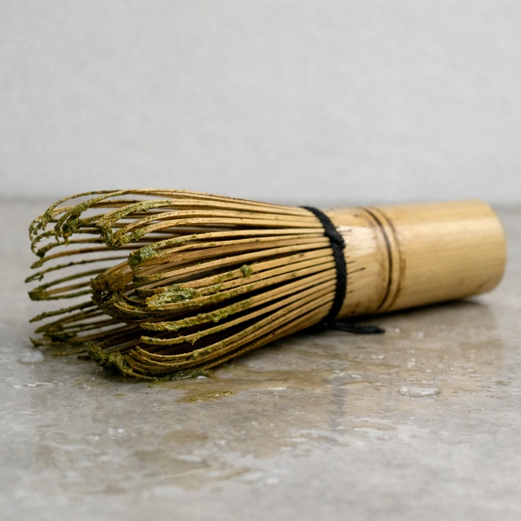 A bamboo matcha whisk lying flat on its side on a wet surface, tines pressed down and slightly bent
