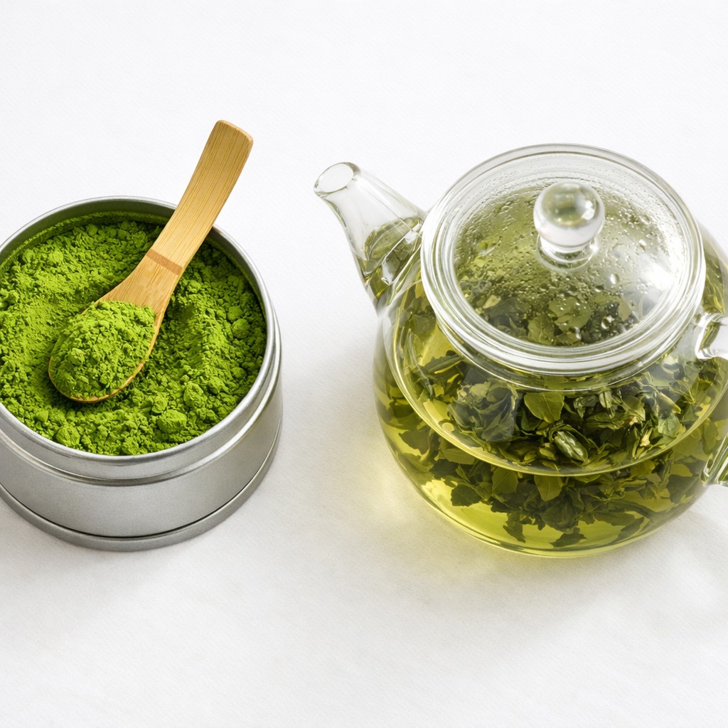 Tin of matcha powder with bamboo scoop next to a glass teapot with steeping green tea leaves